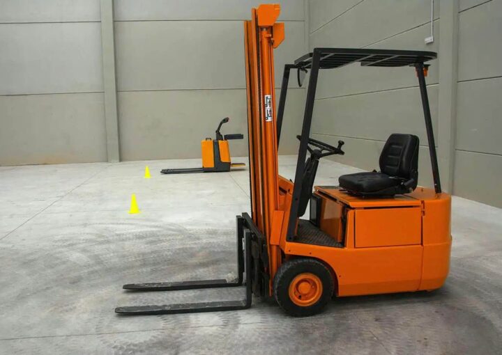 An orange forklift parked in a spacious warehouse with gray walls, showcasing its vertical lifting mechanism and driver seat.