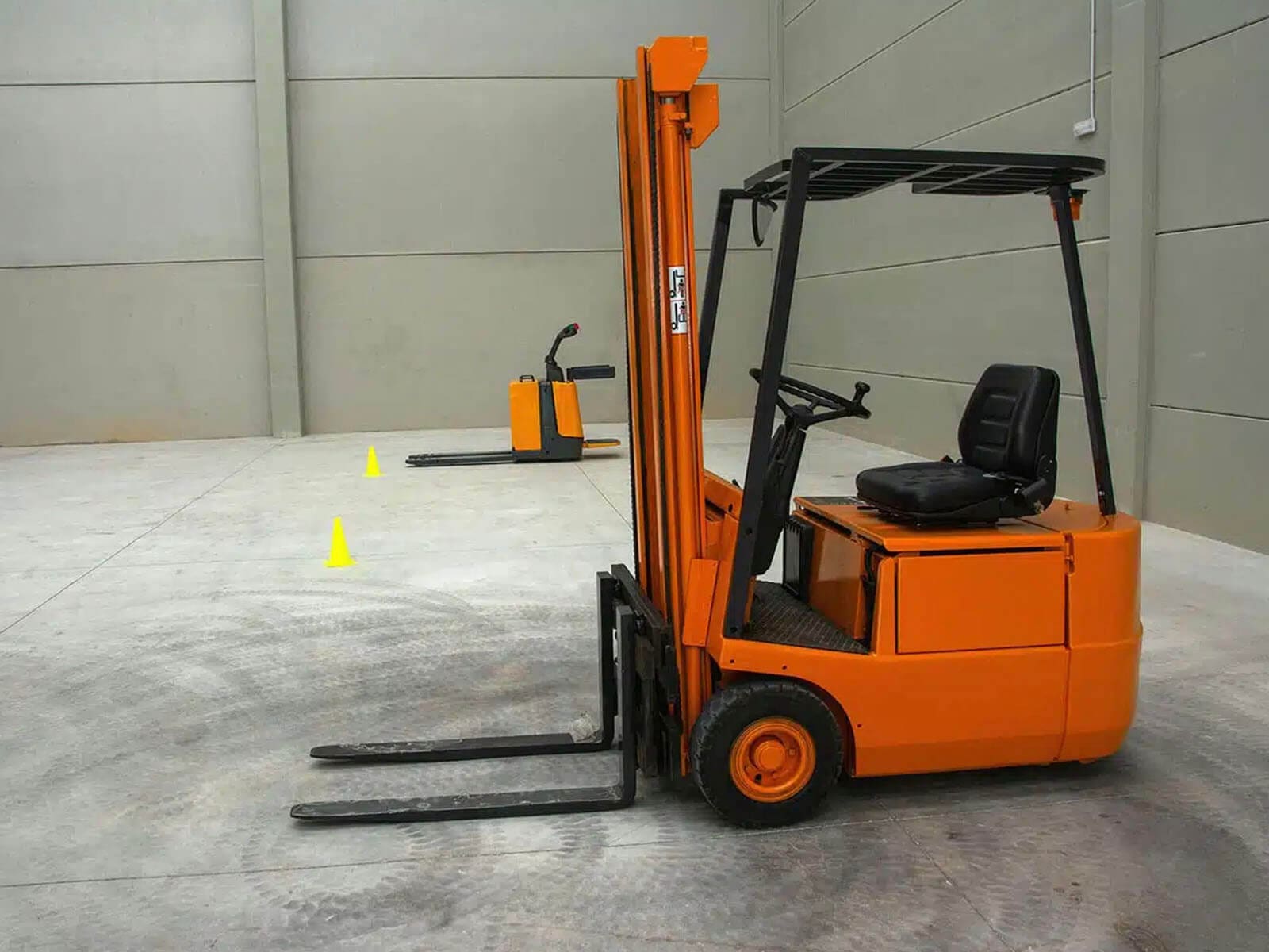 An orange forklift parked in a spacious warehouse with gray walls, showcasing its vertical lifting mechanism and driver seat.