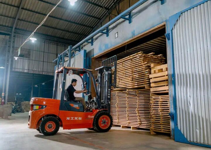 A forklift operator loads stacked wooden pallets inside a warehouse.