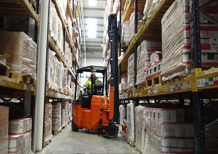 A forklift loaded with supplies navigating through a narrow aisle of a warehouse, surrounded by tall shelves of packaged goods.