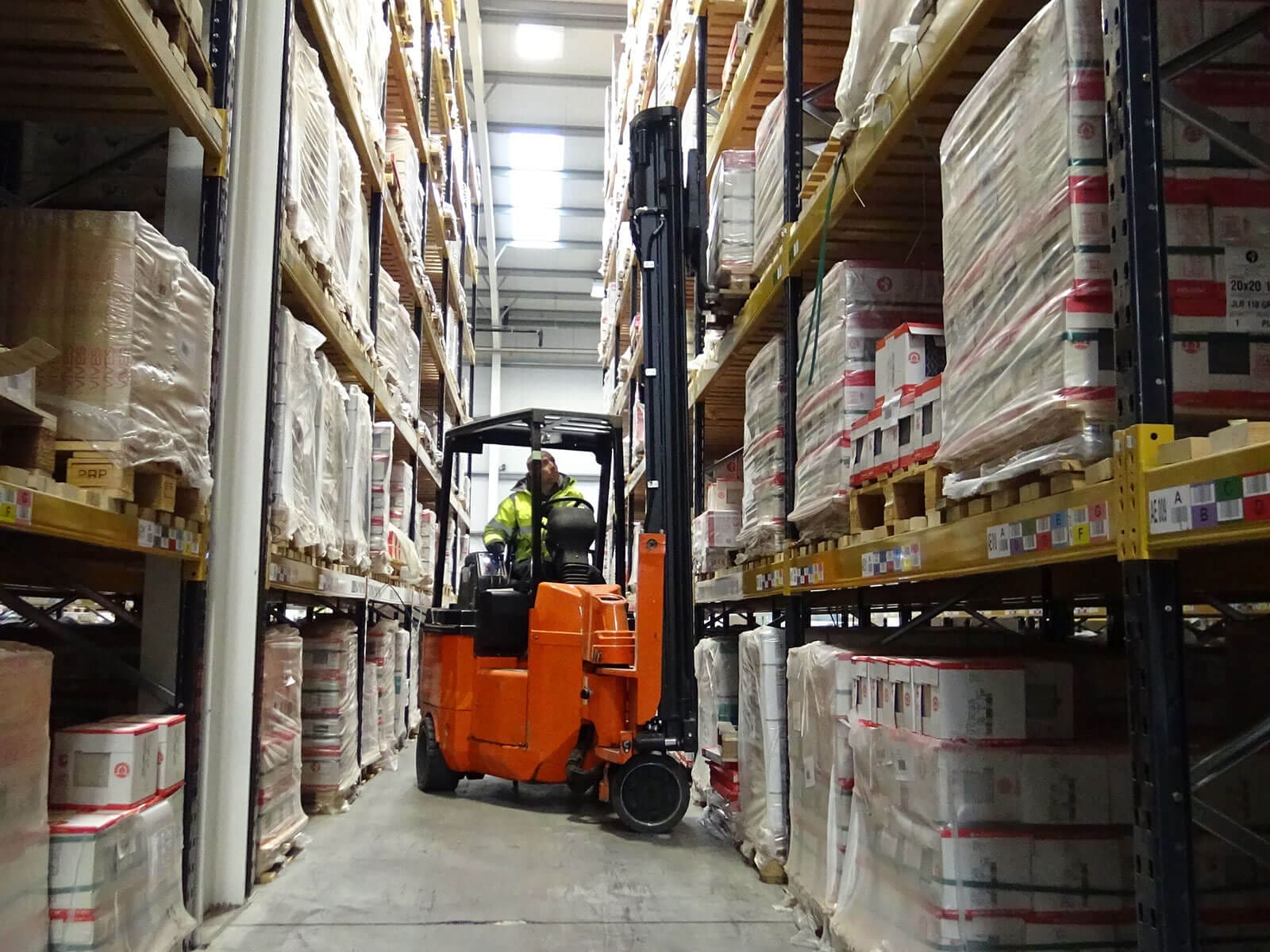 A forklift loaded with supplies navigating through a narrow aisle of a warehouse, surrounded by tall shelves of packaged goods.