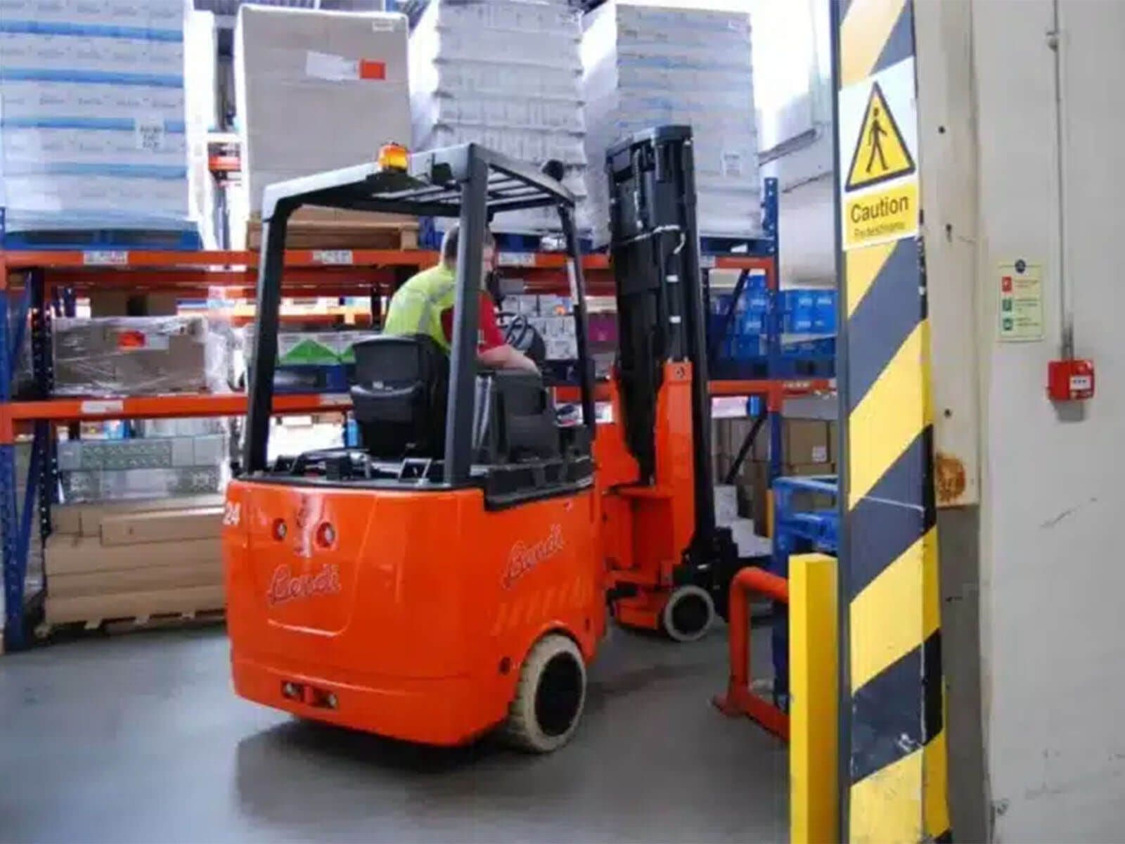 An electric forklift maneuvering in a warehouse aisle filled with stacked pallets and safety signage.